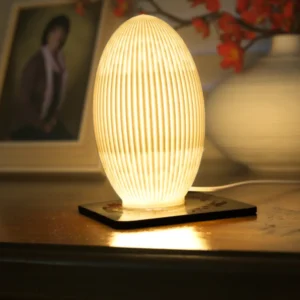 Ribbed 3D printed Japandi egg lamp glowing on a wooden sideboard with photo frame and ceramic vase