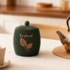 tealeaf-container-tea-setup Forest green ribbed 3D printed tealeaf container styled on a dining table with a glass teapot and a fresh cup of tea.